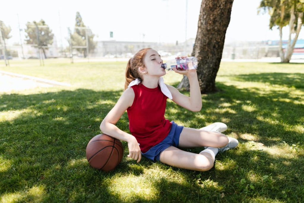 Mädchen sitzt im Schatten und trinkt Wasser nach Sport. Im Artikel: Kinder sollten sich 3 Stunden bewegen?