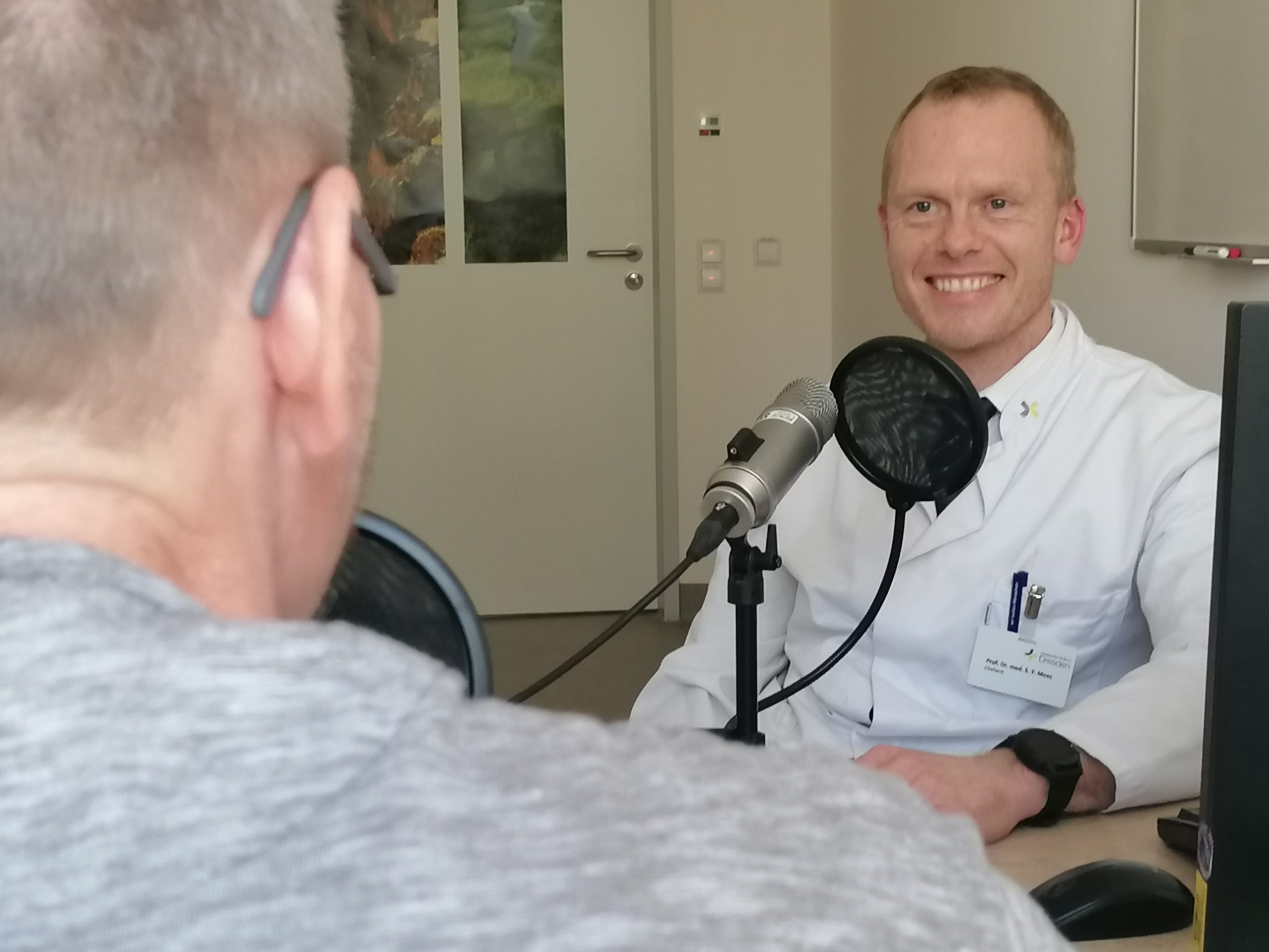 Ein lächelnder Mann im weißen Arztkittel sitzt im Gespräch hinter einem Aufnahmemikrofon
