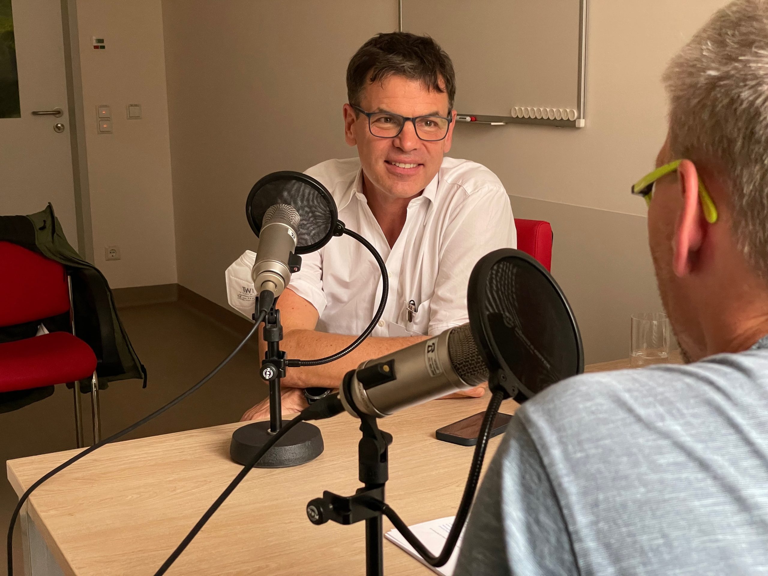 Ein freundlich lächelnder Mann im weißen Arztkittel mit Brille sitzt erzählend hinter einem Mikrofon