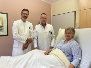 Zwei Ärzte im weißen Arztkittel stehen freundlich lächelnd an einem Patientenbett mit einer Frau im mittleren Alter