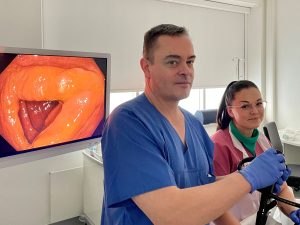 Vor einem großen Bildschirm, auf dem ein Darm von innen zu sehen ist, steht ein Mann im blauen Arztkittel mit einem Endoskop, neben ihm eine junge Frau in einem lila Arztkittel