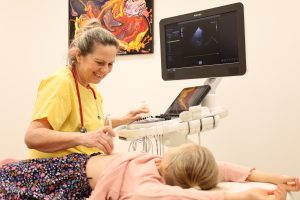 Eine freundlich lachende Frau im gelben Arztkittel untersucht ein auf dem Bauch liegendes Mädchen mit einem Herz-Ultrschall - im Hintergrund ist ein großer Monitor zu sehen