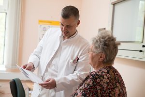 Ein Arzt im weißen Arztkittel erklärt mit Blick auf eine Patientenakte einer älteren Dame einen medizinischen Eingriff